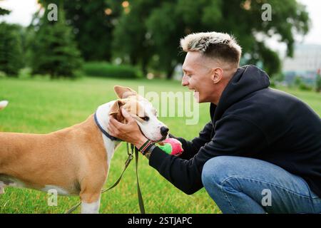 Il giovane con un sorriso in faccia tiene un cane vicino alla museruola che tiene una palla in bocca. Vacanza attiva con un cane. Amore per gli animali bulldo americano Foto Stock