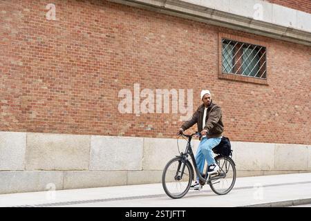 Giovane uomo alla moda che si diverte a fare una piacevole corsa in bicicletta lungo un muro di mattoni in un ambiente urbano Foto Stock