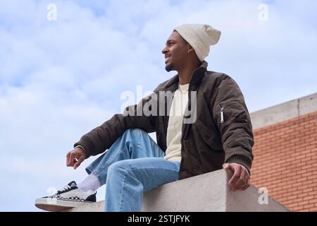 Uomo elegante che indossa un berretto bianco, una giacca marrone e jeans azzurri seduti sul tetto in una giornata nuvolosa Foto Stock