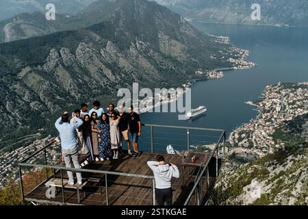 Un gruppo di turisti posa per una foto su una terrazza panoramica che si affaccia sulla splendida baia di Cattaro. Una nave da crociera naviga attraverso le acque calme Foto Stock