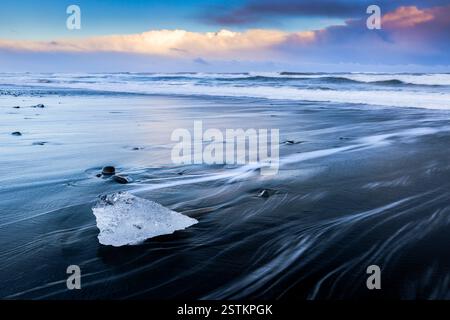 Vista delle onde del mare che scorrono intorno ai blocchi di ghiaccio al tramonto sulla famosa spiaggia Diamond. Jökulsárlón, Diamond Beach, Austurland, Islanda, Nord Europa. Foto Stock