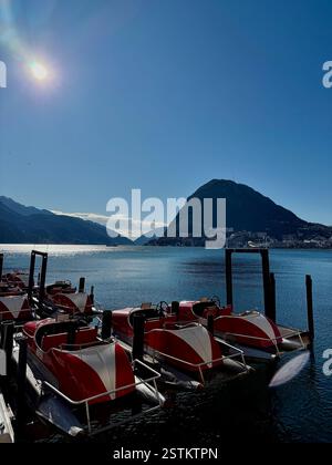 Bellissimo paesaggio del lago di Lugano, Svizzera con piccolo vanto su un molo, acqua blu e montagne sul retro con piccoli edifici sulla riva Foto Stock
