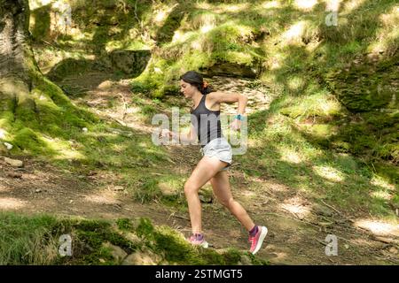 Una giovane donna corre in discesa su un sentiero nella foresta, indossando abbigliamento atletico e scarpe da ginnastica. La luce solare filtra tra gli alberi, evidenziando il muschio verde Foto Stock