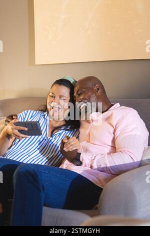 Felice coppia anziana e diversificata seduta sul divano, condividendo momenti di gioia con lo smartphone, a casa. Anziani, felicità, pensionamento, tecnologia, comunicazione, Foto Stock