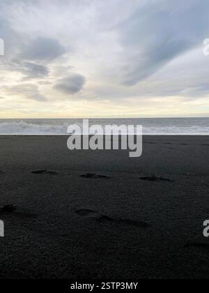 L'immagine cattura una tranquilla scena di spiaggia con sabbia nera, onde dolci e un cielo nuvoloso, evocando un senso di pace e solitudine Foto Stock