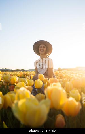Una bella donna con un abito elegante e un cappello di paglia si siede e ride in un vibrante prato giallo tulipano, godendosi la libertà e la bellezza della natura. Foto Stock