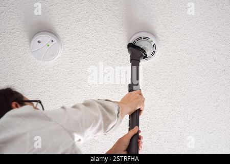 Donna che aspira rilevatori di fumo e monossido di carbonio su un soffitto di una stanza domestica Foto Stock