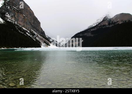 Un paesaggio pittoresco caratterizzato da un lago parzialmente ghiacciato circondato da aspre montagne e foreste sempreverdi, che creano un'atmosfera tranquilla e tranquilla Foto Stock
