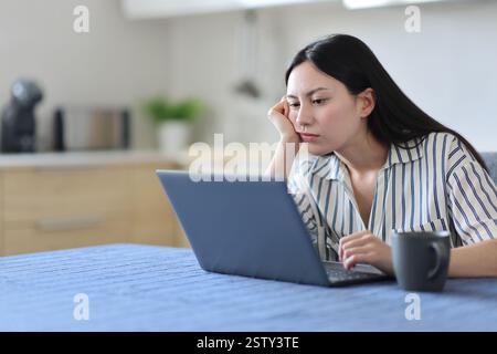 Annoiata donna asiatica che controlla il portatile in cucina a casa Foto Stock