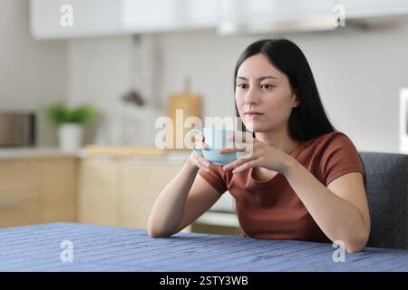 Donna asiatica distratta che beve caffè in cucina a casa Foto Stock