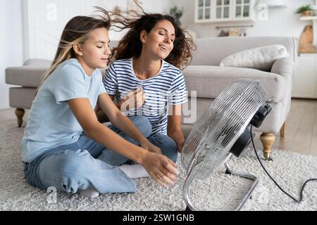 Madre e figlia si raffreddano mentre si siedono sul pavimento di fronte alla ventola elettrica che soffia aria fresca Foto Stock