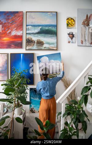 La donna creativa, artista, appende un quadro moderno finito sulla parete in uno studio d'arte domestico con piante d'appartamento. Foto Stock