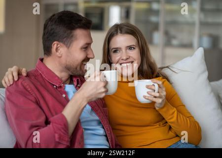Amata coppia uomo donna che passa un fine settimana pigro beve un caffè seduto sul divano a casa, divertitevi a parlare. Foto Stock