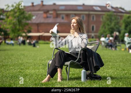 Una donna di mezza età che si prende una pausa sul prato urbano si siede a piedi nudi e salta i documenti per rinfrescarsi. Foto Stock