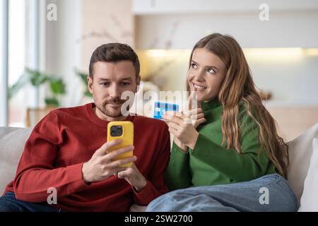 Coppie sposate felici sposi che fanno acquisti online tramite Internet utilizzando lo smartphone, seduti sul divano. Foto Stock