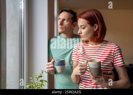 Preoccupazione seria giovane coppia della famiglia in piedi insieme dalla finestra a casa che beve il caffè di mattina Foto Stock