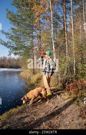 Donna con caccia al cane vizsla che cammina nella foresta vicino allo stagno guardando attraverso il binocolo sugli uccelli. Foto Stock
