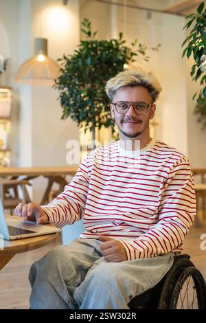 Un uomo con ritratti disabili, libero professionista su sedia a rotelle, siede in un bar adattivo con computer portatile. Foto Stock