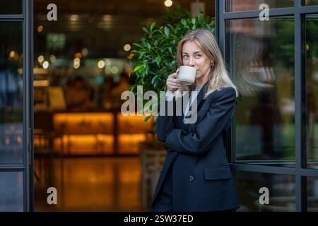 Una donna d'affari di mezza età, fiduciosa e contenta, beve caffè mattutino all'entrata della caffetteria Foto Stock