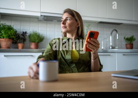 Una donna premurosa siede in cucina con una tazza che regge lo smartphone e che sogna ad occhi aperti la finestra. Foto Stock