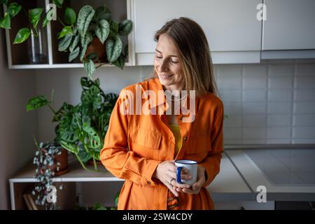 Donna gioiosa sorridente in piedi in una cucina verde con una tazza di caffè che prepara la giornata al mattino Foto Stock