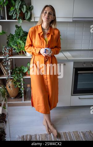 Donna felice rilassata in piedi nella cucina di casa tenendo la tazza guardando la telecamera circondata dalle piante d'appartamento Foto Stock