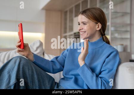 Una donna felice effettua una piacevole videochiamata con lo smartphone seduto sul divano di casa sorridendo guardando lo schermo Foto Stock