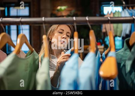 Donna interessata a fare shopping, scegli l'abbigliamento in un negozio alla moda accanto a un rack con le camicie Foto Stock