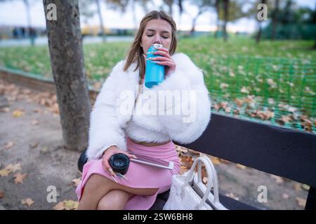 Giovane donna che beve da una bottiglia riutilizzabile nel parco mentre è seduta sulla panchina, godendo la giornata autunnale Foto Stock