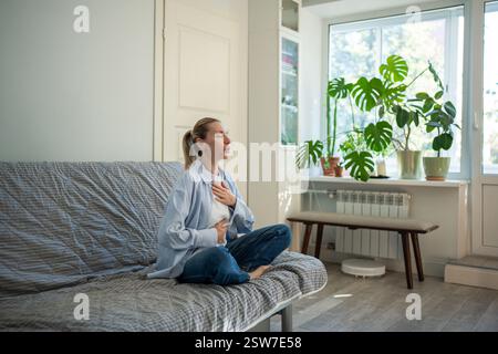 Meditazione a casa di una donna di mezza età seduta nel loto posa respiro profondo toccando il petto e la pancia Foto Stock