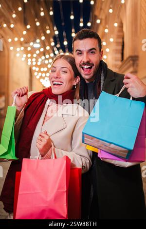 Verticale. Coppia gioiosa che celebra una spedizione di shopping di successo tenendo in mano le valigie e condividendo un momento delizioso pieno di risate e felicità durante un'affascinante gita serale insieme. Foto di alta qualità Foto Stock