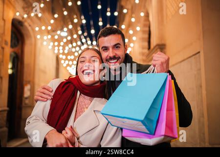 Coppia gioiosa che celebra una spedizione di shopping di successo tenendo in mano borse colorate e condividendo un momento delizioso pieno di risate e felicità durante un'affascinante gita serale insieme. Foto di alta qualità Foto Stock