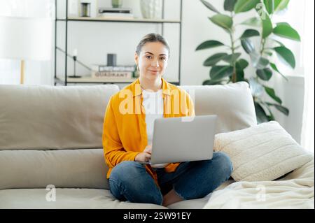 Giovane donna caucasica sicura e affascinante con una camicia gialla, seduta su un comodo divano con un laptop e sorridente davanti alla fotocamera in una casa moderna e luminosa. Concetto di lavoro online, tecnologia e produttività Foto Stock