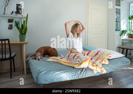 Si è svegliata positivamente la mattina, una sbadigliante sdraiata si siede a letto con un cane ottimista la giornata di inizio. Foto Stock