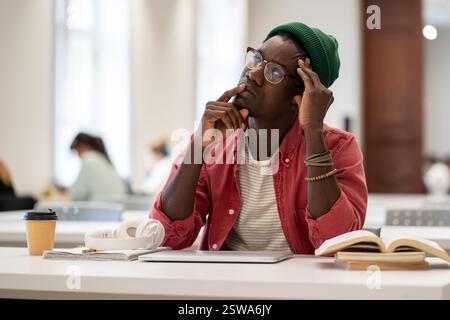 Un ragazzo afroamericano perplesso ha perso nei pensieri mentre studiava nella biblioteca dell'università Foto Stock