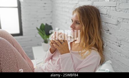 La giovane donna bionda si gode una tazza di caffè mentre si rilassa nella sua camera da letto con pareti in mattoni bianchi e pigiama rosa. Foto Stock