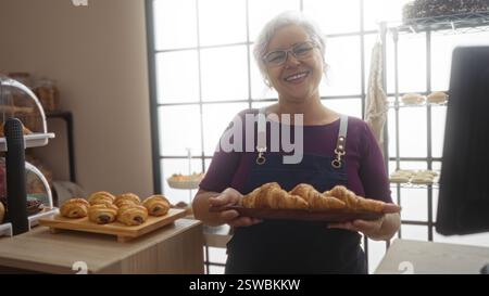 Donna anziana ispanica con i capelli grigi sorridente mentre tiene i croissant in un accogliente negozio di panetteria interno. Foto Stock