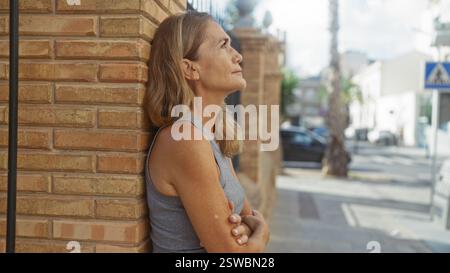 Donna con le braccia incrociate si appoggia a un muro urbano in mattoni all'aperto, contemplando la vita cittadina, incarnando la bellezza matura e la calma sicurezza. Foto Stock