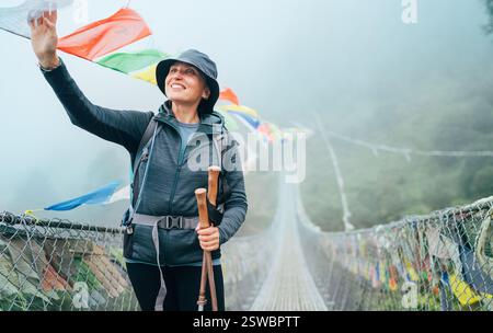 Giovane donna sorridente che attraversa il canyon sul ponte sospeso e toccando le bandiere di preghiera tibetane multicolori incernierate sulla gola. Percorso di arrampicata sulla vetta di Mera Foto Stock