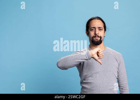 Giovane persona insoddisfatta che mostra il simbolo del pollice in basso in studio, esprimendo la sua negazione e rifiuto su sfondo blu. Un uomo serio scontento che posa con un simbolo antipatico. Foto Stock