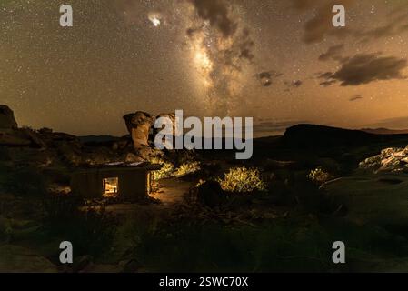 Cielo notturno della via Lattea su capanna di Adobe illuminata e formazioni rocciose nel paesaggio del deserto Foto Stock