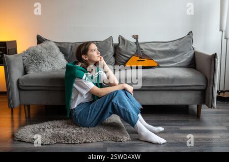 Donna premurosa e ispirata seduta su un tappeto e sognando, mentre fai una pausa nelle prove della balalaika a casa Foto Stock