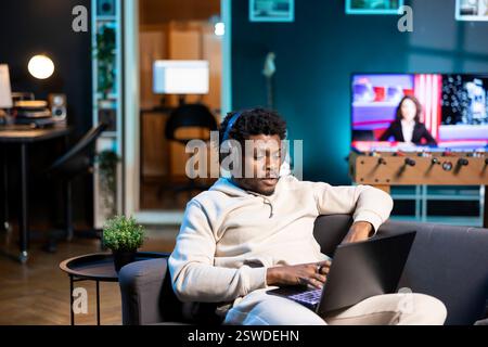 Dipendente remoto che ascolta podcast audio mentre lavora da casa e lavora in multitasking. Un libero professionista indossa le cuffie, digita sulla tastiera del laptop nell'elegante ufficio personale dell'appartamento Foto Stock