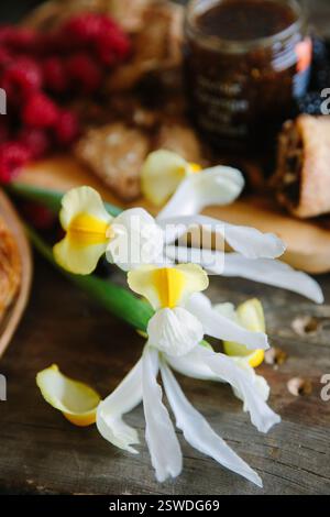 Orchidee bianche e gialle fresche si trovano accanto a un assortimento di pasticcini e frutta su un caldo tavolo di legno. Foto Stock
