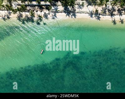 Acqua verde smeraldo con fondali marini visibili e una piccola barca da pesca vicino alla riva, fiancheggiata da ombre di palme. Koh Samui, Thailandia. Foto Stock