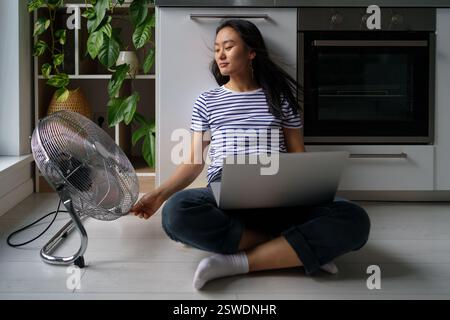 Felice ragazza asiatica sorridente freelancer seduto vicino a un ventilatore elettrico che lavora da casa in onda di calore Foto Stock