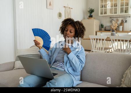 Una donna africana stanca che sventola il ventilatore in mano soffre di calore durante la giornata di lavoro con il laptop sul divano di casa Foto Stock