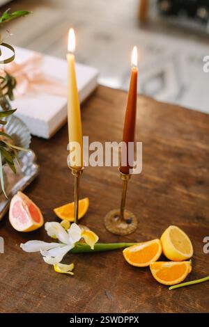 Le candele illuminano un tavolo di legno con agrumi e un fiore, creando un'atmosfera calda e invitante. Foto Stock