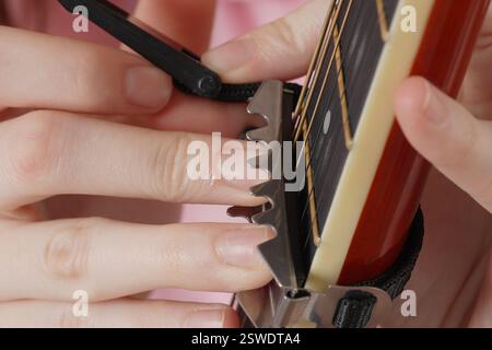Un primo piano estremo di dita femminili collega il capotasto alla chitarra acustica a sei corde. Capodastro o capo tasto è un dispositivo che i chitarristi usano sul collo Foto Stock
