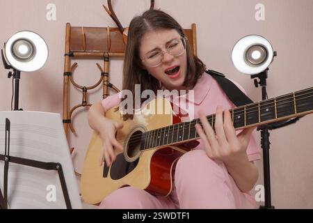 La giovane donna con gli occhiali da vista suona la chitarra acustica a sei corde e canta la canzone con tanta emozione Foto Stock